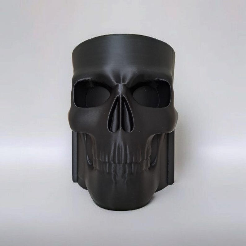 May include: A black skull-shaped planter with a wide opening. The planter has detailed facial features, including eye sockets, a nose cavity, and a jawline. The skull is made of a matte black material and is suitable for indoor or outdoor use.
