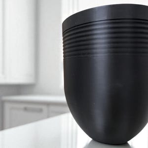 May include: A matte black, rounded container with a textured, ribbed band near the top. The container has a smooth, curved body and sits on a reflective surface. The background is a blurred white interior.