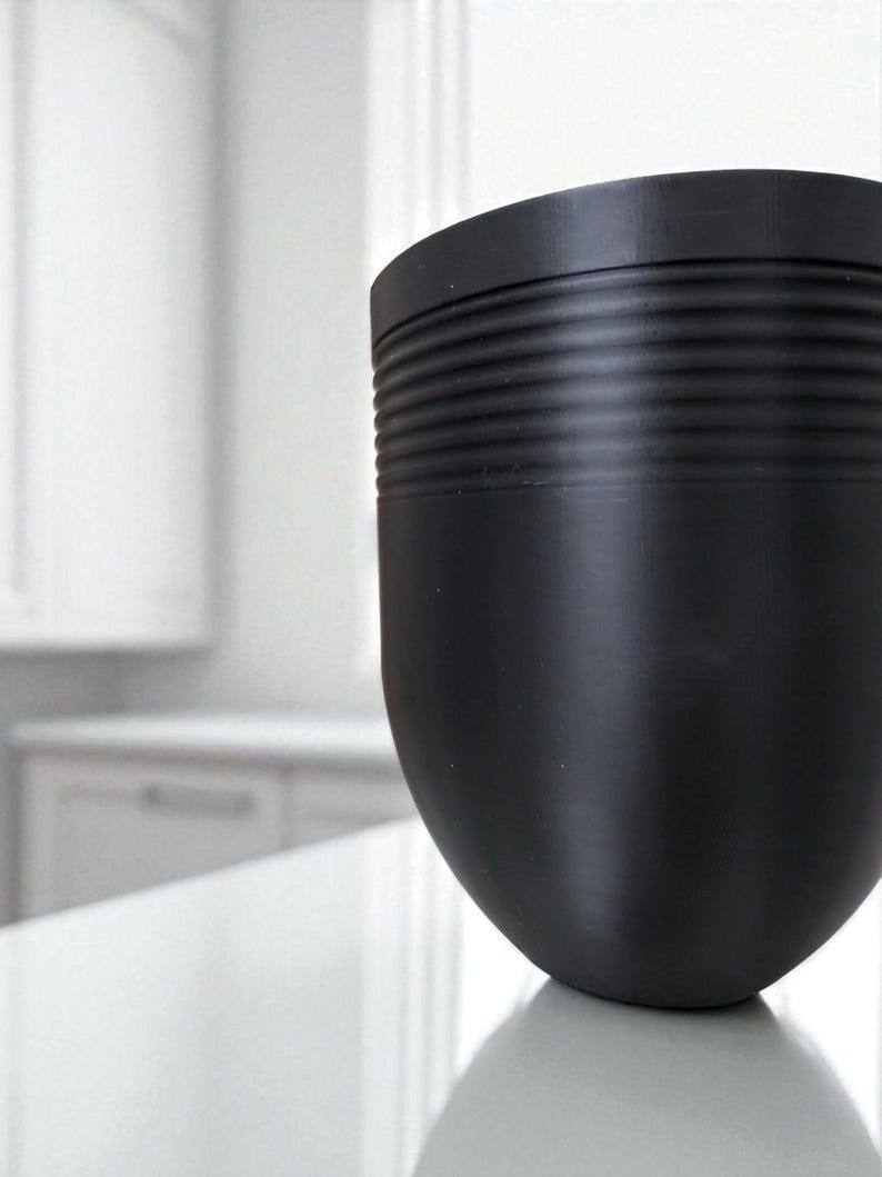 May include: A matte black, rounded container with a textured, ribbed band near the top. The container has a smooth, curved body and sits on a reflective surface. The background is a blurred white interior.