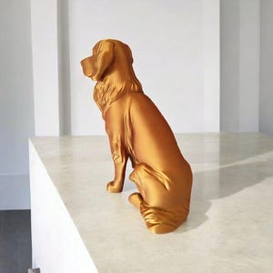 May include: A golden-colored dog statue in a seated position. The sculpture has a smooth, reflective surface and detailed features, including the dog's ears, fur, and paws. The statue is placed on a white surface.