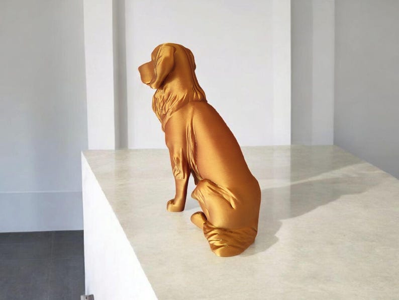 May include: A golden-colored dog statue in a seated position. The sculpture has a smooth, reflective surface and detailed features, including the dog's ears, fur, and paws. The statue is placed on a white surface.