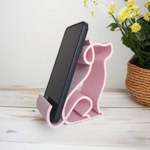May include: A pink phone stand in the shape of a dog, holding a black smartphone. The stand is made of a solid material and has a smooth finish. The dog-shaped stand is on a white wooden surface.