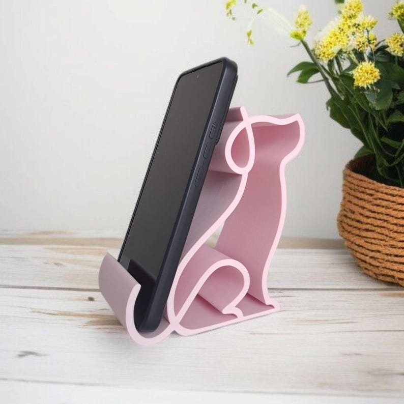 May include: A pink phone stand in the shape of a dog, holding a black smartphone. The stand is made of a solid material and has a smooth finish. The dog-shaped stand is on a white wooden surface.