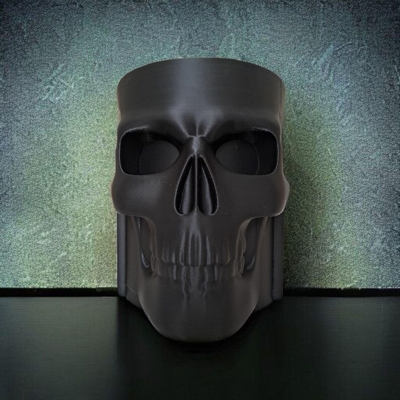 May include: A black skull-shaped decorative container. The container is made of a matte black material and features detailed facial features, including eye sockets, a nose cavity, and teeth. The container is designed to stand upright.