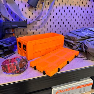 May include: Bright orange rectangular storage container with a matching lid, and a red and black camouflage watch. The container has black latches and is sitting on a shelf. Other items are visible in the background.