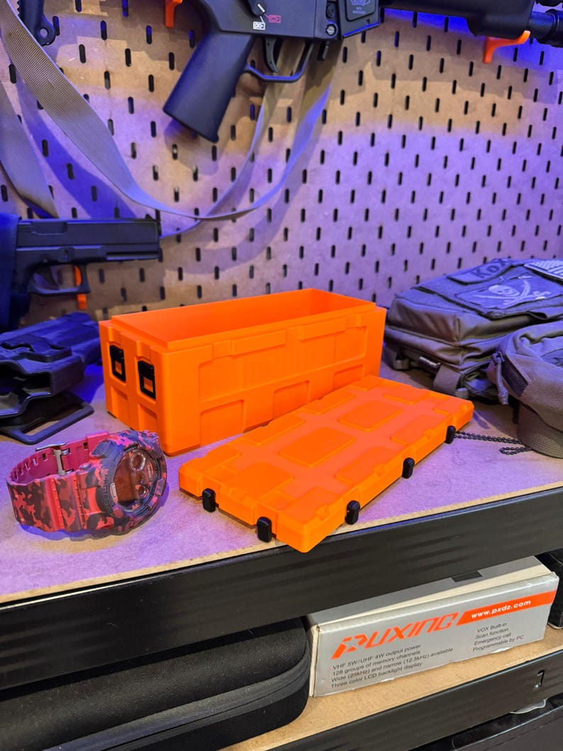 May include: Bright orange rectangular storage container with a matching lid, and a red and black camouflage watch. The container has black latches and is sitting on a shelf. Other items are visible in the background.