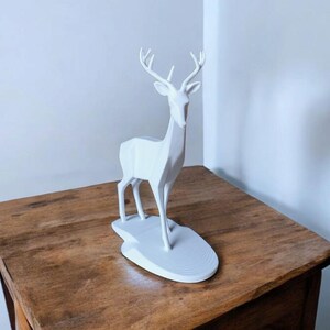 Puede incluir: Una escultura de ciervo geom&eacute;trico blanco con cuernos se alza sobre una base blanca. El ciervo est&aacute; colocado sobre una mesa de madera. La escultura tiene un dise&ntilde;o moderno y minimalista, adecuado para la decoraci&oacute;n del hogar.