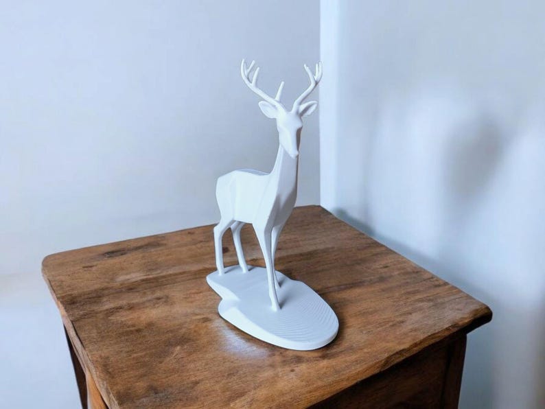 Puede incluir: Una escultura de ciervo geom&eacute;trico blanco con cuernos se alza sobre una base blanca. El ciervo est&aacute; colocado sobre una mesa de madera. La escultura tiene un dise&ntilde;o moderno y minimalista, adecuado para la decoraci&oacute;n del hogar.