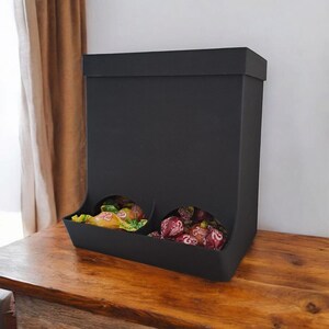 May include: A black candy dispenser with two compartments, each filled with wrapped candies. The dispenser is rectangular with a flat top and a curved opening at the bottom of each compartment. It sits on a wooden surface.