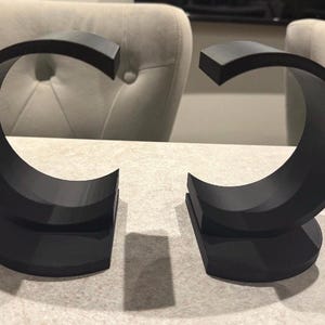 May include: Two black, C-shaped bookends. Each bookend has a flat, semi-circular base and a curved upper portion. The bookends are made of a solid, matte material and are designed to hold books upright.