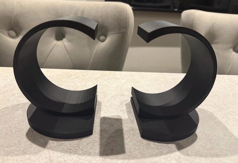 May include: Two black, C-shaped bookends. Each bookend has a flat, semi-circular base and a curved upper portion. The bookends are made of a solid, matte material and are designed to hold books upright.