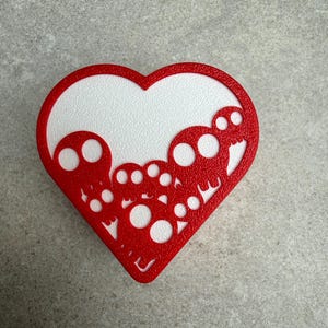 May include: A red heart-shaped decoration with a white textured interior. The red border features a cut-out design of skulls. The decoration is placed on a gray surface.