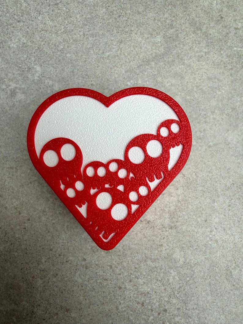 May include: A red heart-shaped decoration with a white textured interior. The red border features a cut-out design of skulls. The decoration is placed on a gray surface.