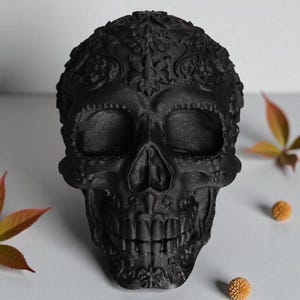 May include: A black decorative skull with intricate floral patterns. The skull has detailed facial features, including eye sockets, a nose cavity, and teeth. The skull is a dark matte black color and is surrounded by fall foliage.