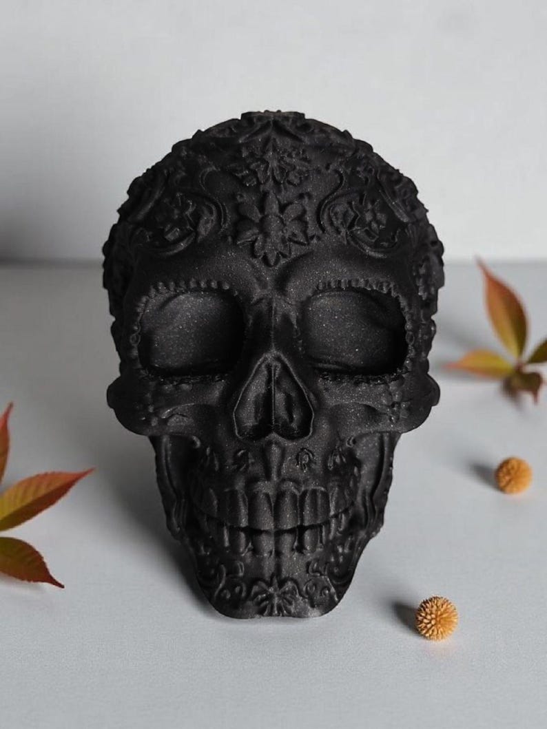 May include: A black decorative skull with intricate floral patterns. The skull has detailed facial features, including eye sockets, a nose cavity, and teeth. The skull is a dark matte black color and is surrounded by fall foliage.