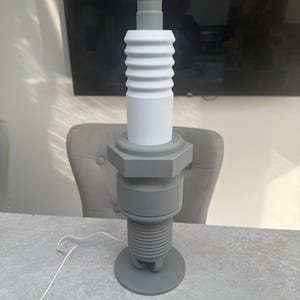 May include: A gray and white decorative lamp shaped like a spark plug. The lamp has a cylindrical design with a rounded top and a threaded base. A white cord extends from the base.
