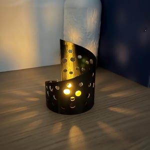 May include: A black metal candle holder with a semi-circular design and numerous circular cutouts. The candle holder is illuminated by a warm, yellow light, casting shadows on the wooden surface. A white, textured decorative object is in the background.