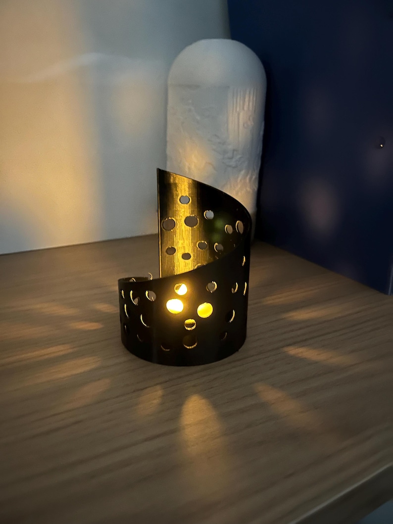 May include: A black metal candle holder with a semi-circular design and numerous circular cutouts. The candle holder is illuminated by a warm, yellow light, casting shadows on the wooden surface. A white, textured decorative object is in the background.