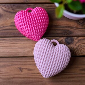 May include: Two heart-shaped items with a knitted texture, one bright pink and the other light pink, are shown on a wooden surface. Each heart has a small loop at the top. A potted plant with pink flowers is in the background.