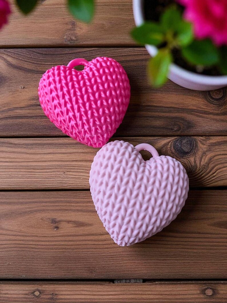 May include: Two heart-shaped items with a knitted texture, one bright pink and the other light pink, are shown on a wooden surface. Each heart has a small loop at the top. A potted plant with pink flowers is in the background.