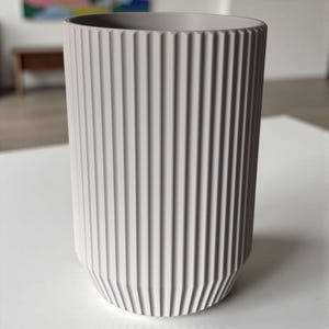 May include: A tall, cylindrical, light gray ceramic vase with vertical ribbed detailing. The vase has a slightly tapered base and a smooth, unadorned rim. The background includes abstract art and a white surface.