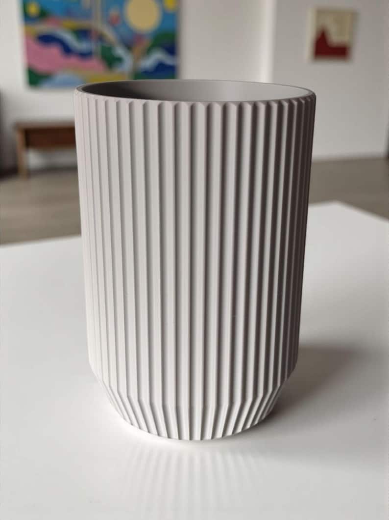 May include: A tall, cylindrical, light gray ceramic vase with vertical ribbed detailing. The vase has a slightly tapered base and a smooth, unadorned rim. The background includes abstract art and a white surface.