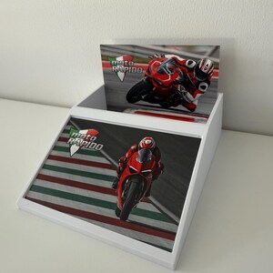 May include: White display stand with images of a red motorcycle racing on a track. The stand has the text "moto RAPIDO" in green and white. The images show a motorcycle racer in red gear.
