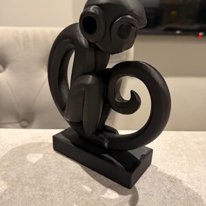 Puede incluir: Escultura negra de un mono sentado, con una cola enroscada y grandes ojos redondos. El mono est&aacute; posado sobre una base rectangular. La escultura est&aacute; hecha de un material oscuro y s&oacute;lido.