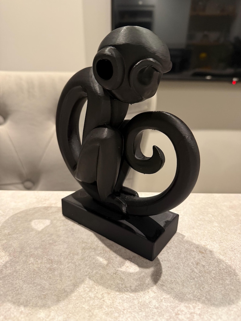 Puede incluir: Escultura negra de un mono sentado, con una cola enroscada y grandes ojos redondos. El mono est&aacute; posado sobre una base rectangular. La escultura est&aacute; hecha de un material oscuro y s&oacute;lido.
