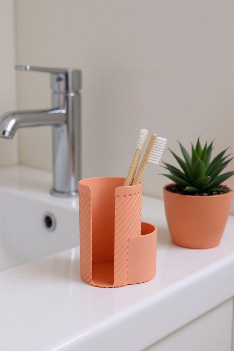 May include: A peach-colored toothbrush holder with two bamboo toothbrushes. The holder has a textured design and a curved section. A small potted succulent plant sits to the right. The scene is set in a bathroom.
