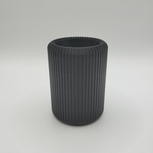 May include: A cylindrical, dark gray container with vertical ribbed detailing. The top rim has a textured, ridged design. The container is made of a solid material and stands on a white surface.