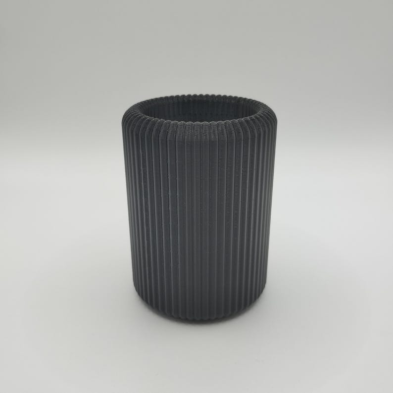 May include: A cylindrical, dark gray container with vertical ribbed detailing. The top rim has a textured, ridged design. The container is made of a solid material and stands on a white surface.