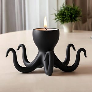 May include: A black octopus-shaped candle holder with a lit candle. The candle holder has eight curved tentacles and a round base. The candle flame is yellow and orange. The holder is on a light-coloured surface.