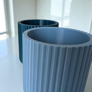 May include: Two cylindrical planters with vertical ridged detailing. One is a muted teal color, and the other is a light blue-grey. The planters are made of a solid material and have a textured surface. The planters are suitable for indoor or outdoor use.