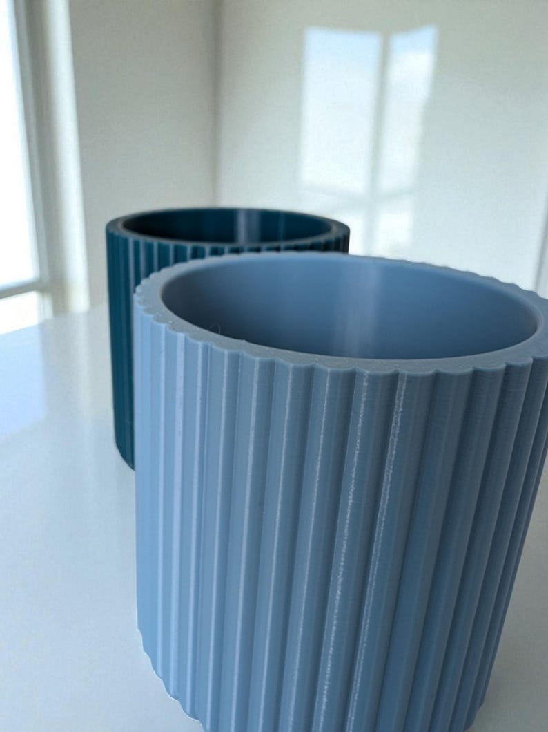 May include: Two cylindrical planters with vertical ridged detailing. One is a muted teal color, and the other is a light blue-grey. The planters are made of a solid material and have a textured surface. The planters are suitable for indoor or outdoor use.