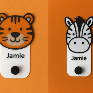 May include: Two animal-themed name tags. One features a tiger face with orange and black stripes, the other a zebra face with black and white stripes. Each tag has the name "Jamie" printed on a white rectangular base with a black button.