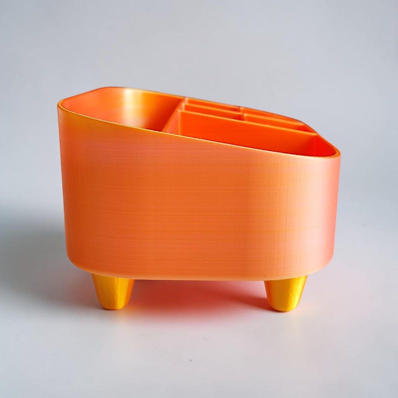 May include: An orange organizer with a gradient effect, featuring multiple compartments for storage. The organizer has a rounded rectangular shape and is supported by three yellow legs. The design is modern and functional, suitable for desk organization.