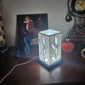 May include: A lit, square lamp with a white shade featuring black leaf designs. The lamp sits on a dark base and is plugged in. A painting of a fox and a decorative clock are in the background.