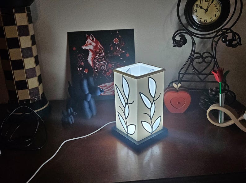 May include: A lit, square lamp with a white shade featuring black leaf designs. The lamp sits on a dark base and is plugged in. A painting of a fox and a decorative clock are in the background.