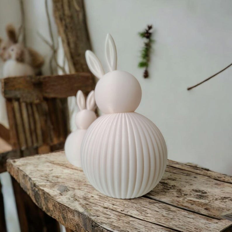 May include: Three white ceramic bunny figurines. The largest bunny has a spherical head and a ribbed, spherical body. The smaller bunnies are in the background. The bunnies are on a rustic wooden surface.