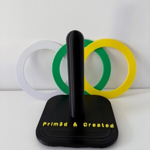 May include: A black paper towel holder with a vertical post and a square base. The base has the text "Prim3d & Created" in yellow. Behind the holder are three interlocking rings in white, green, and yellow.