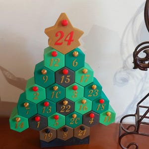 May include: A green, gold, and brown Christmas tree-shaped advent calendar. The tree is made of hexagonal compartments, each numbered from 1 to 24. The star on top has the number 24. Red and gold accents decorate the tree.