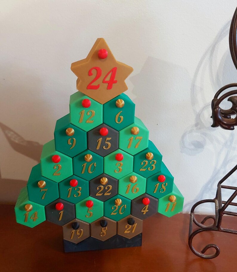 May include: A green, gold, and brown Christmas tree-shaped advent calendar. The tree is made of hexagonal compartments, each numbered from 1 to 24. The star on top has the number 24. Red and gold accents decorate the tree.