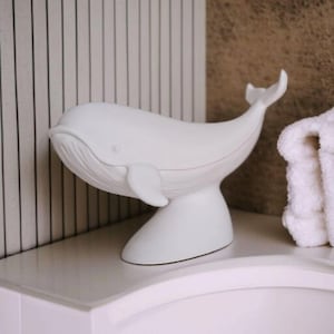 K&ouml;nnte beinhalten: Eine wei&szlig;e Wal-Figur aus Keramik mit geschwungenem K&ouml;rper, Flossen und erhobenem Schwanz. Der Wal steht auf einem Sockel. Der Hintergrund umfasst eine wei&szlig;-schwarz gestreifte Wand und ein wei&szlig;es Handtuch.