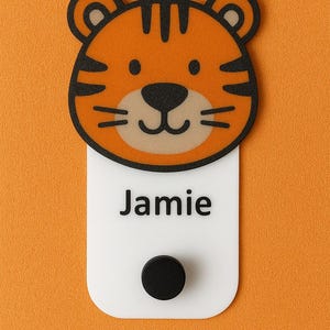 May include: A tiger-faced name tag with the name "Jamie" printed in black on a white rectangular base. The tiger face is orange with black stripes and a black nose. A black circular magnet is at the bottom.