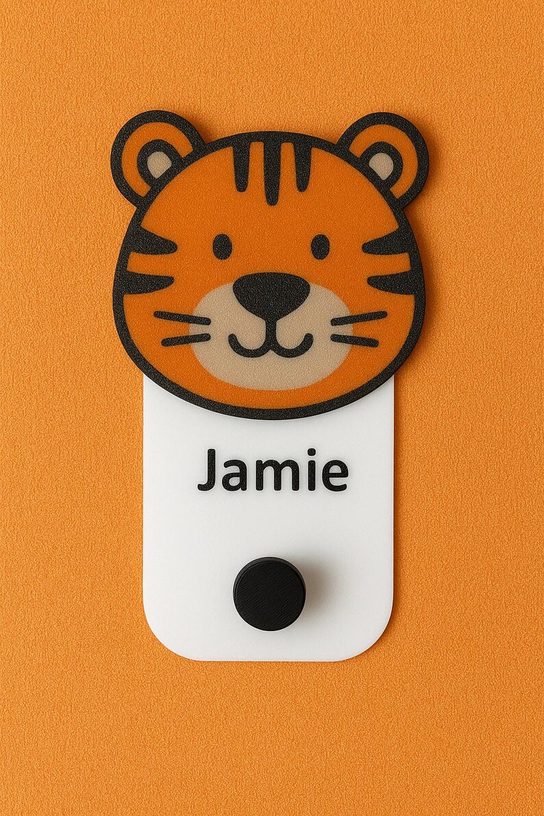 May include: A tiger-faced name tag with the name "Jamie" printed in black on a white rectangular base. The tiger face is orange with black stripes and a black nose. A black circular magnet is at the bottom.