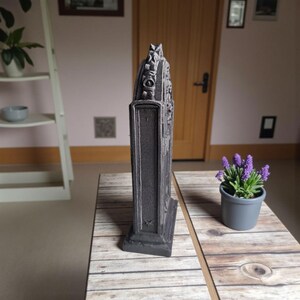 May include: A dark gray, rectangular decorative object with ornate detailing. The object has a textured surface and stands upright on a wooden table. A small potted plant with purple flowers sits nearby.