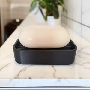 May include: A light-colored bar of soap rests on a black soap dish with a slatted design. The soap dish is rectangular and sits on a white countertop with a marble pattern. The background is blurred.