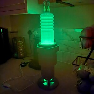 May include: A green glowing lamp shaped like a spark plug. The lamp has a cylindrical body with ribbed detailing and a hexagonal base. The lamp is lit with a bright green light, and the base has a circular design.