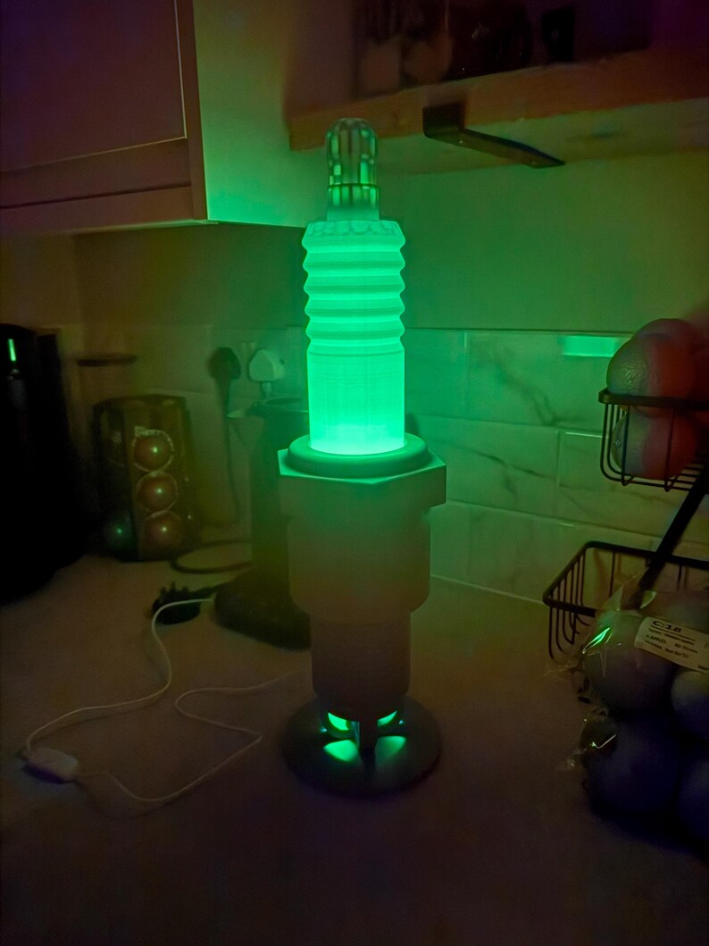 May include: A green glowing lamp shaped like a spark plug. The lamp has a cylindrical body with ribbed detailing and a hexagonal base. The lamp is lit with a bright green light, and the base has a circular design.